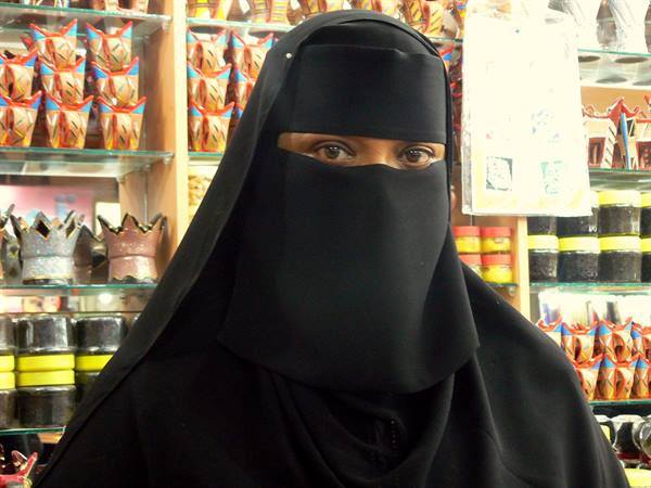 Woman in black niqab inside a perfume and incense shop, shelves full of products