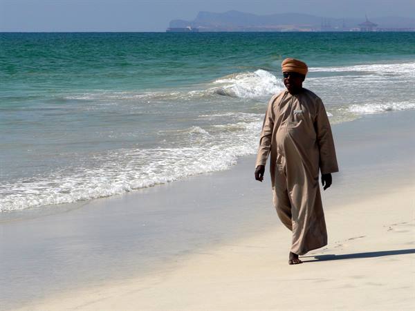 Man in beige dishdasha and turban walks barefoot on sand near waves
