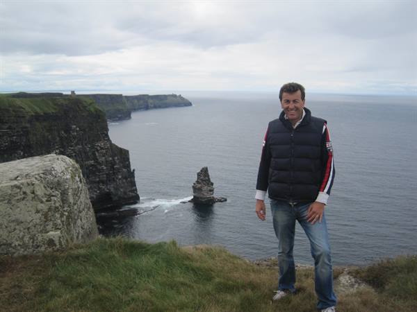 Smiling man in down jacket at the Cliffs of Moher, cliffs plunging into the ocean