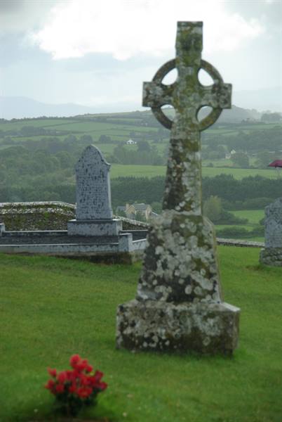 Moss-covered Celtic cross in green cemetery, misty rolling hills of Cashel behind