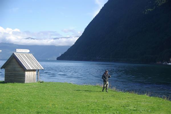 Angler with rod beside wooden hut on grass, fjord surrounded by mountains