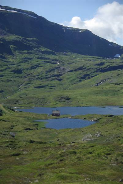 Red wooden cabin by a green lake among grassy snowy mountains
