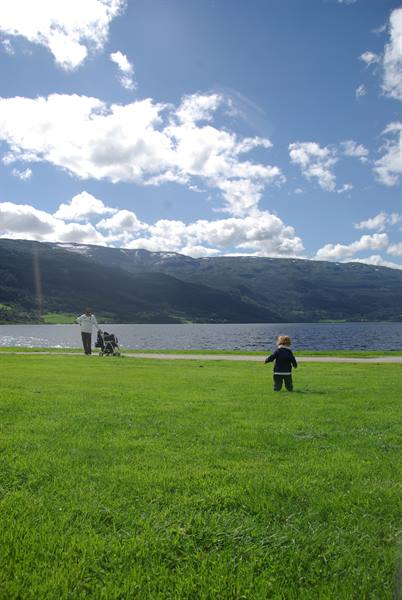 Child on green lawn by the lake, parent with stroller, mountains and clouds
