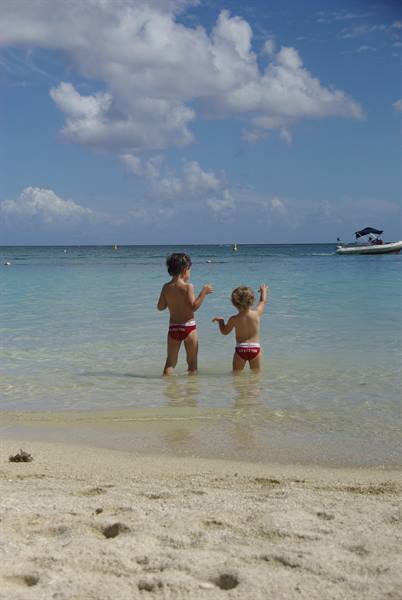 Two children in red swimsuits play in turquoise water by the sandy beach