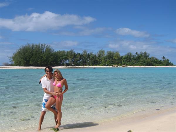 Couple standing in turquoise water on white sand, green islet with pines behind