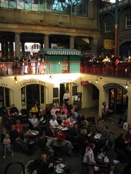 crowd indoors, balcony railings above a green kiosk, people seated at tables under yellow signs