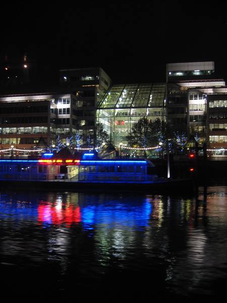 Blue and red illuminated ferry reflects on river at night among glass buildings