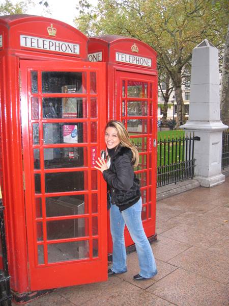 Smiling woman poses beside red telephone booths in a square with trees