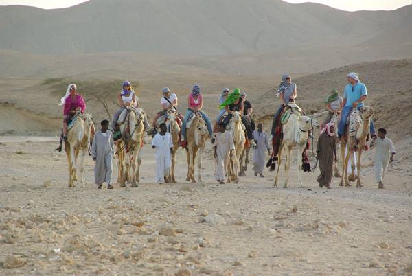 Tourists riding camels in a line led by robed guides in the desert