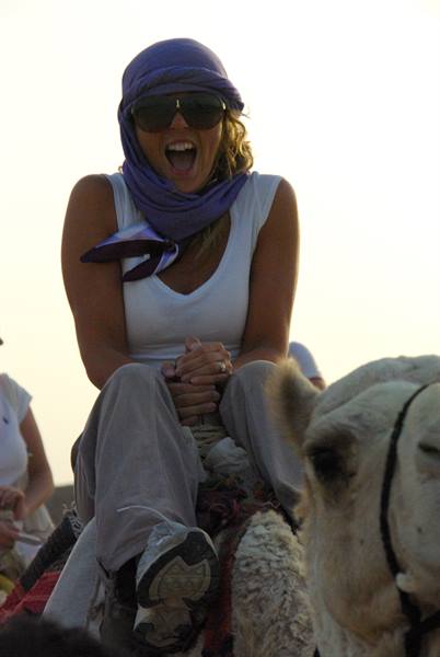 Woman in purple headscarf and sunglasses laughs riding a camel