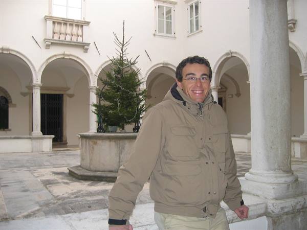 Smiling man in a stone courtyard with arched colonnade and central well