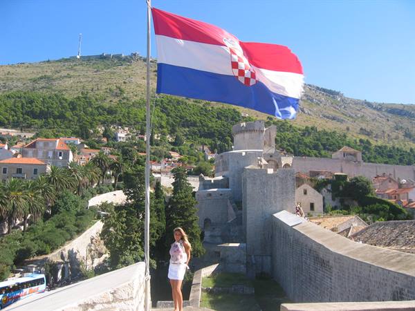 Croatian flag waving over Dubrovnik walls, woman posing, green hill behind