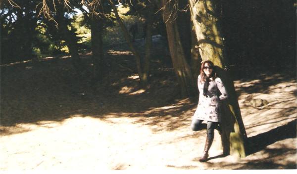 Woman in coat, scarf and boots leaning against a tree trunk in a wood