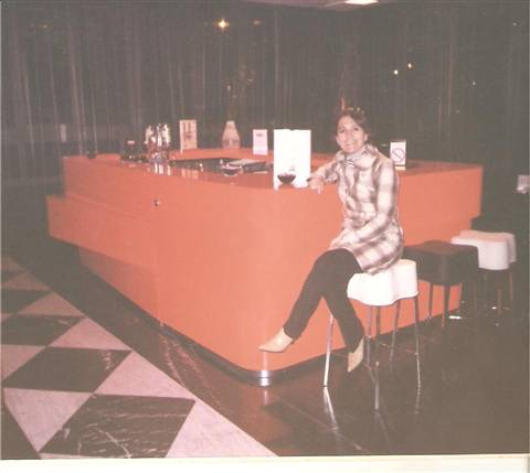 Woman sitting on a stool beside an orange bar counter, checkered floor
