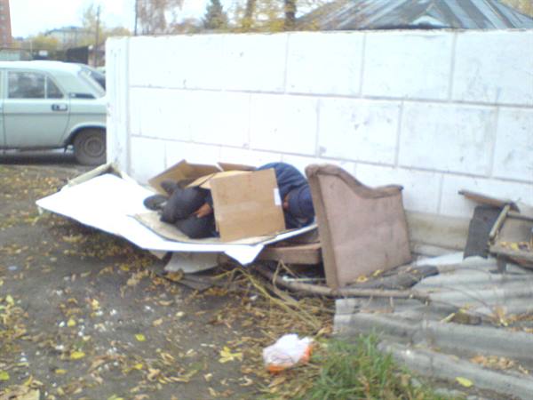 Man sleeps inside cardboard by a white brick wall, vintage car nearby