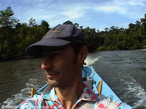 Man in cap and printed shirt riding a wooden boat along a jungle river