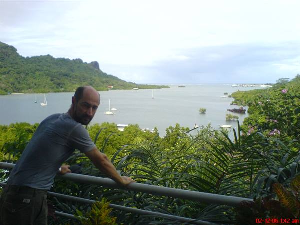 Man leaning on railing over bay with sailboats and green hills