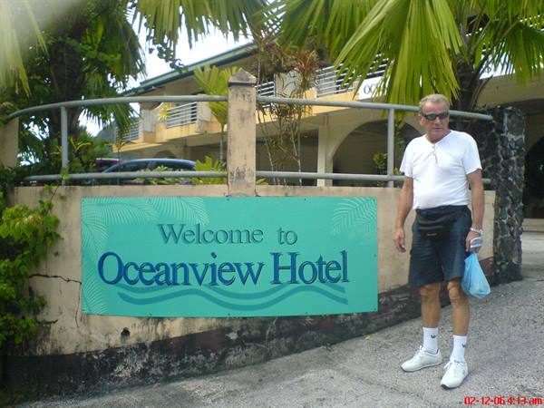 Man with bag beside the Welcome to Oceanview Hotel sign among palm trees