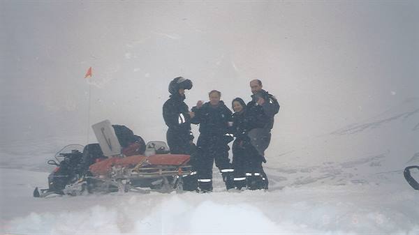 Four people in snowsuits beside a snowmobile with a stretcher in foggy snow