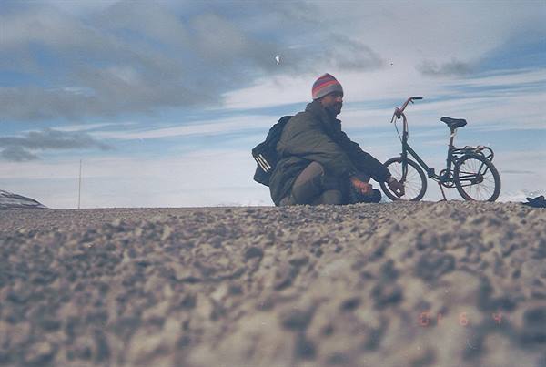 Man in beanie with backpack beside a folding bike on gravel under cloudy sky