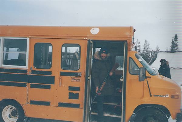 Man in parka steps out of a yellow bus in snow among spruce trees