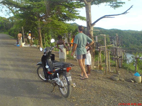 Tourist browses wooden souvenir stalls beside dirt road, scooter parked nearby