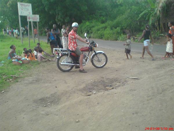 Helmeted tourist stopped on red motorbike on dirt road, people sitting on grass