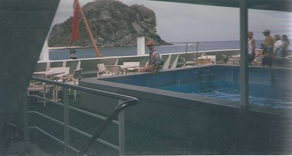 Man in red hat beside ship's pool with a rocky islet behind