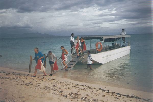 Tourists disembark from a white boat onto a sandy beach under cloudy skies