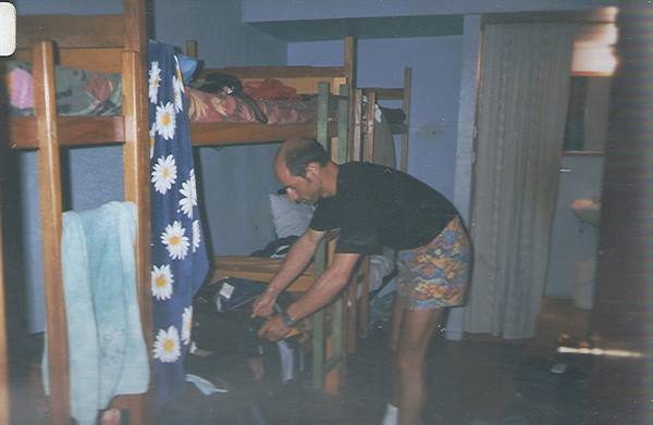 Man in black tank top and colorful shorts packs his backpack among wooden hostel bunk beds