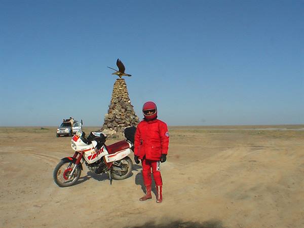 Rider in red suit next to motorcycle before stone cairn topped with eagle