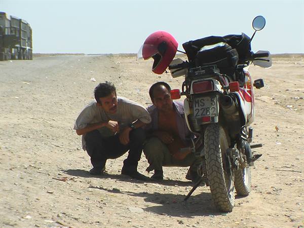 Two men crouching beside a motorcycle with red helmet on desert road