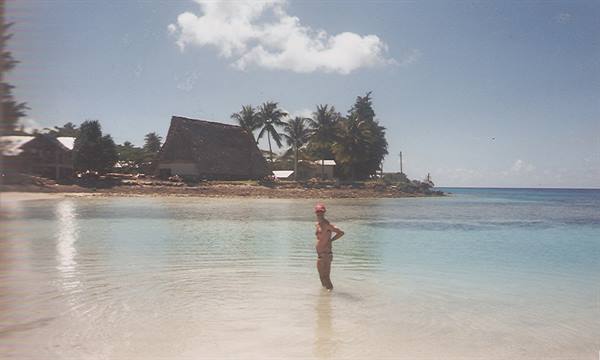 Woman in swimsuit wading in clear water, thatched hut and palms ashore