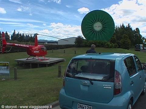 Blue car parked near a red helicopter on a grassy platform