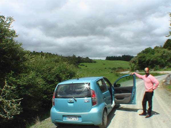 Man in pink shirt opens the door of a blue car on a gravel road