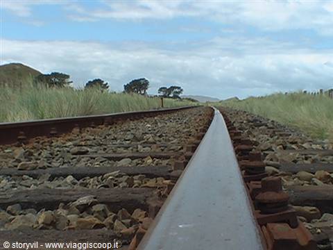 Rusty rail track curving through tall grass and sandy dunes under cloudy sky