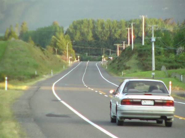 White car driving on a rural asphalt road between grassy hills and trees