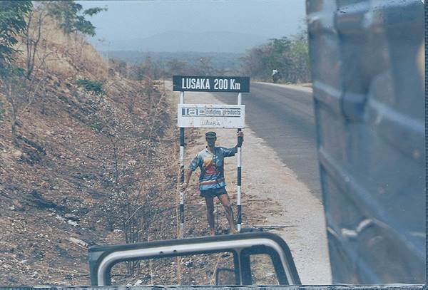 Man beside Lusaka 200 km sign on dusty roadside