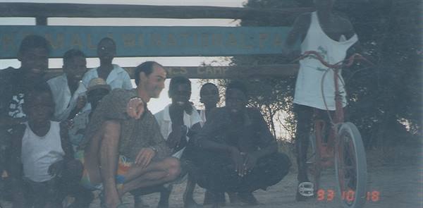 Group of men and boys with a bicycle in front of a green sign on a dirt road