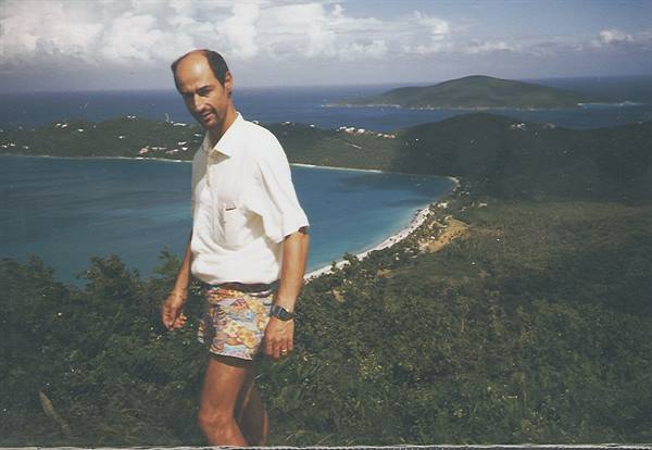 Man in white shirt and floral shorts on green hillside above turquoise bay