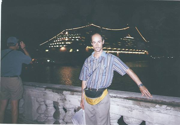 Man in striped shirt leaning on stone balustrade, illuminated cruise ship at night