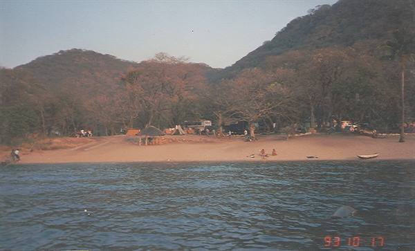 Sandy beach with campsite among trees, green mountains behind, calm waters
