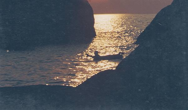 Swimmer silhouetted between rocky cliffs, sparkling sea under low sun