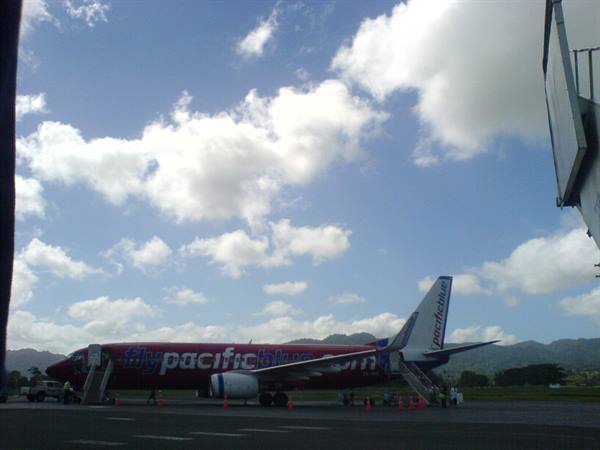 Red Pacific Blue plane parked on tarmac with boarding stairs and green mountains behind