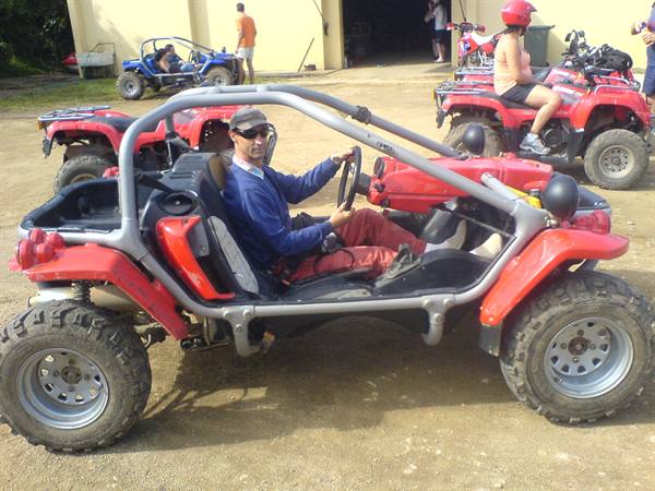Man in cap and sunglasses driving red buggy with gray roll cage on dirt ground