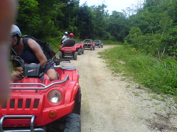 Red ATV convoy on a dirt road through tropical jungle, riders wearing helmets