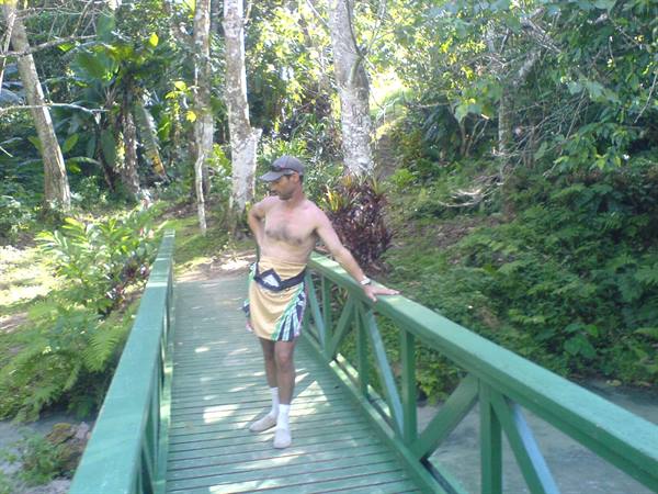 Man in cap and sarong leaning on the green railing of a wooden jungle bridge