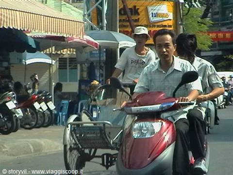 Cycle rickshaws and red scooters weaving through busy city street traffic