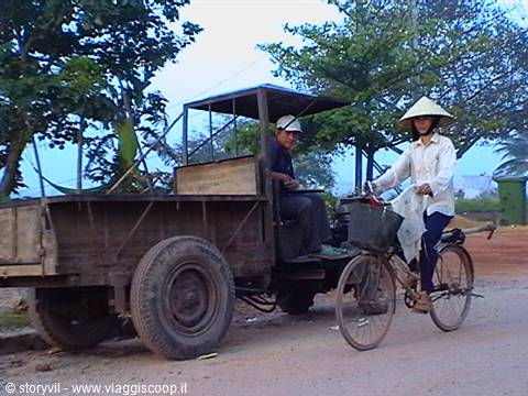 Woman in straw hat cycling beside an old wooden truck on a dirt road