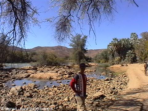 Boy standing on rocks by a river, palm trees and mountains under blue sky